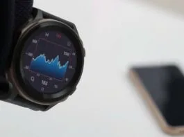 A digital watch screen showing graph data, with a phone visible in the distance.